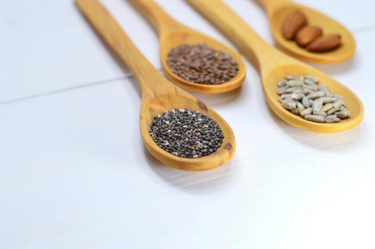 A variety of seeds and nuts in wooden spoons displayed on a white surface.